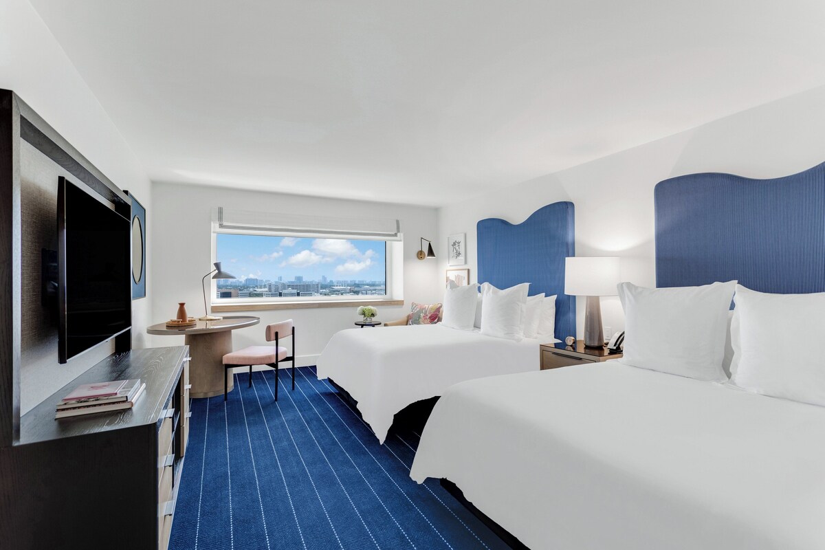 A hotel room with two beds, a desk, a TV, a window with a city view, and a blue carpet.