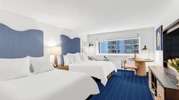 Ein Hotelzimmer mit zwei Betten, einem Schreibtisch, einem Stuhl, einem Fenster mit Blick auf Gebäude und einem Flachbildfernseher.