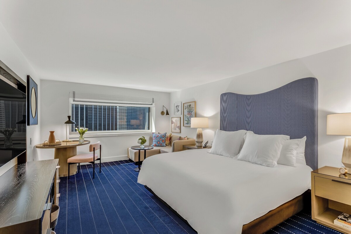 A hotel room with a large bed, a desk, a chair, a small table with a vase, and a window with blinds.