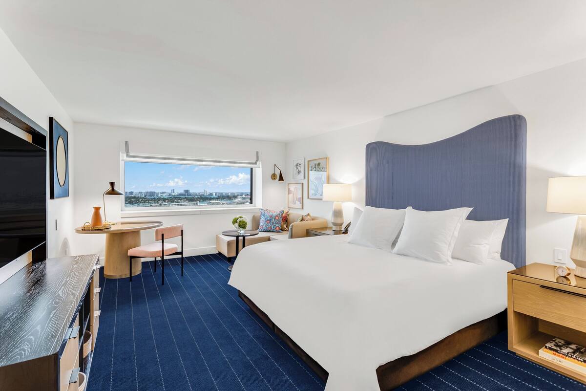A hotel room with a large bed, a desk with a chair, a small table with a vase, and a window with a city view.