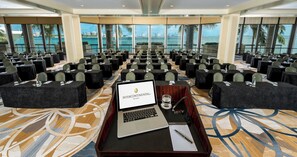 A conference room with rows of tables and chairs, a laptop on a podium, and a view of the ocean through large windows.