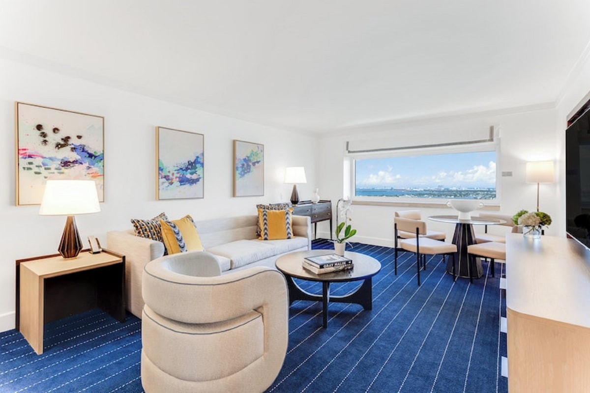 A modern hotel room with a sofa, armchairs, a coffee table, and a TV. There are artworks on the wall, a window with a view, and a desk with a lamp.