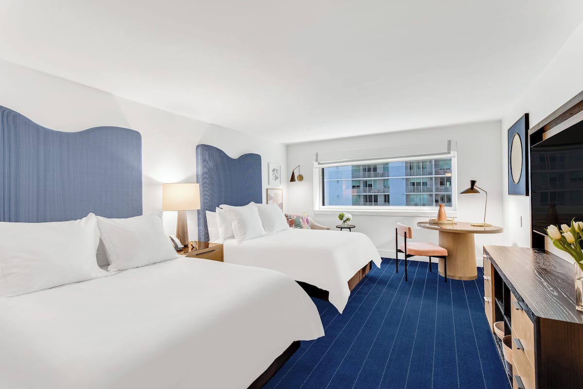 A hotel room with two beds, a desk, a chair, a small table, a lamp, and a window with a city view.