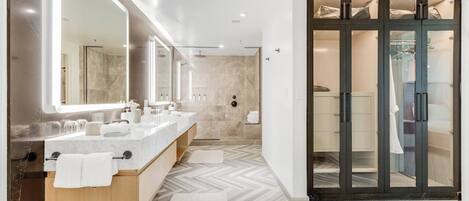 A modern bathroom with a large mirror, two sinks, a shower, and a walk-in closet.