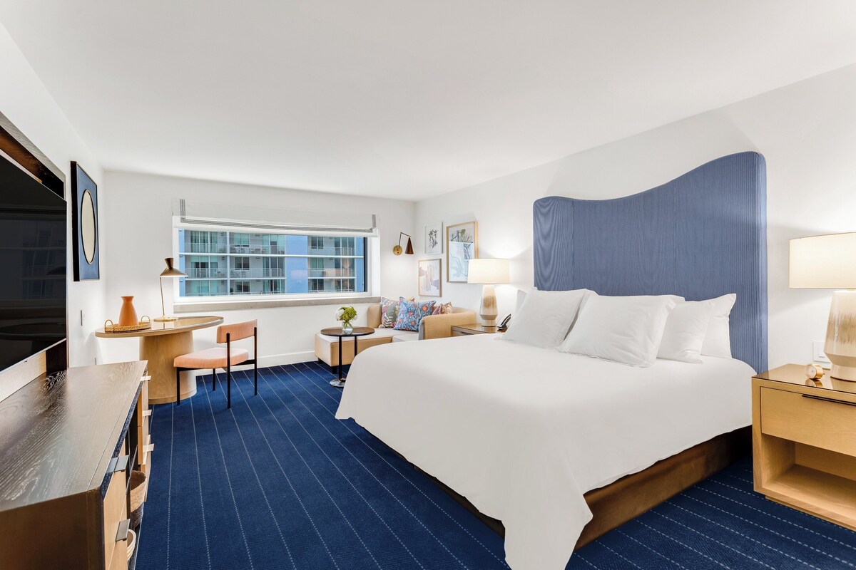 A hotel room with a large bed, a desk, a chair, a window, and a view of a building outside.
