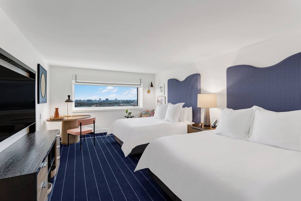 A hotel room with two beds, a desk, a chair, a TV, a window with a city view, and a blue-striped carpet.