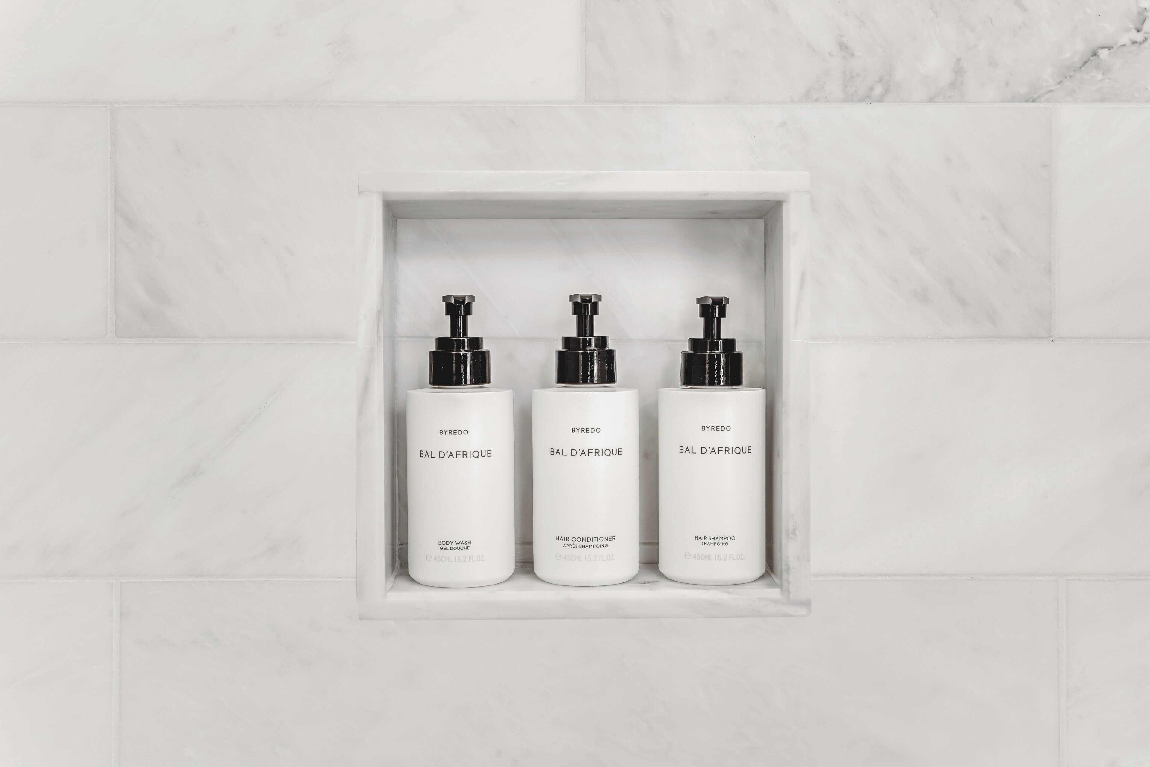 Three white bottles of Byredo Bal D'Afrique products in a marble-tiled alcove.