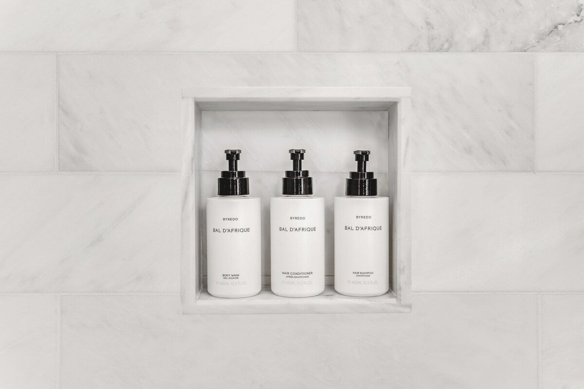 Three white bottles of Byredo Bal D'Afrique products in a marble-tiled alcove.