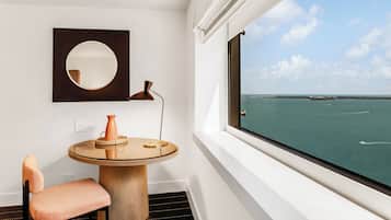 A room with a round table, a chair, a mirror, and a view of the sea through a window.