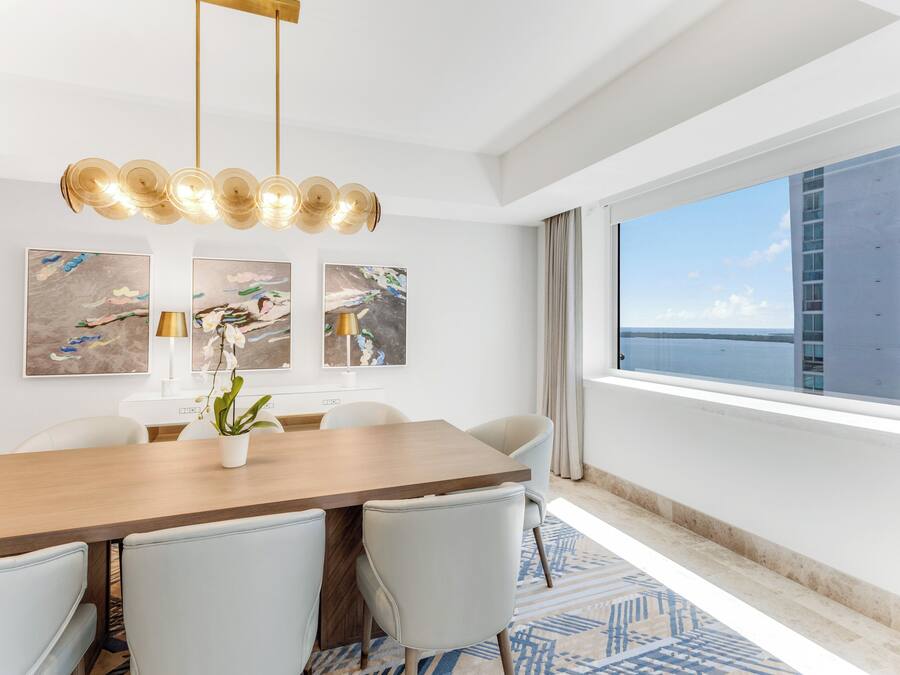A modern dining area with a wooden table, white chairs, and a potted orchid. There are abstract paintings on the wall and a large window with a view of the city.