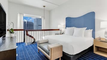 A hotel room with a large bed, a desk, a chair, a small table, a nightstand, a lamp, a potted plant, and a view of the city skyline through a window.
