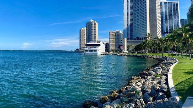 A waterfront cityscape with high-rise buildings, a yacht docked near the shore, and a well-maintained grassy area with palm trees.