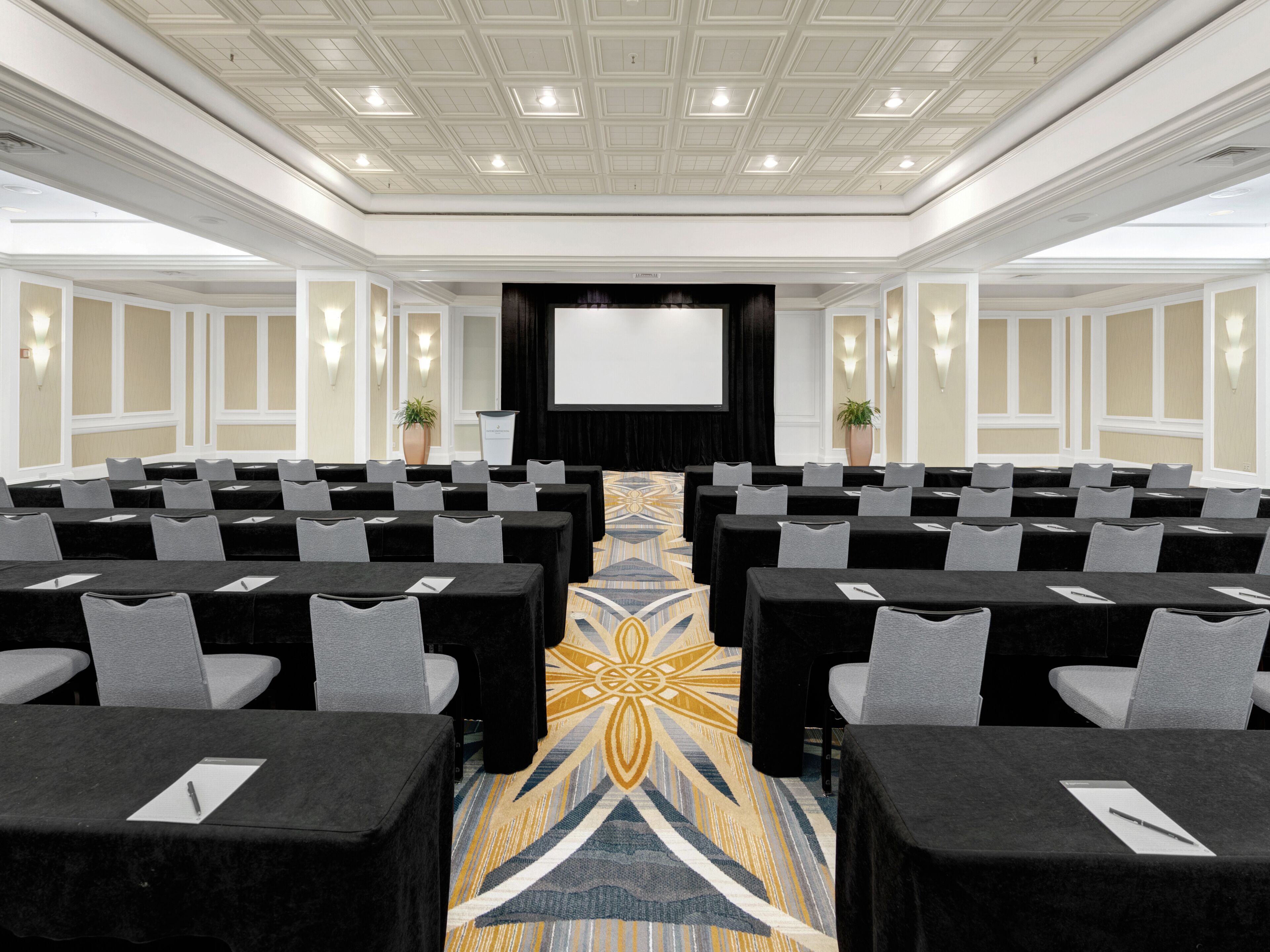 Une salle de conférence avec des rangées de tables et de chaises, un grand écran à l’avant et un tapis à motifs.