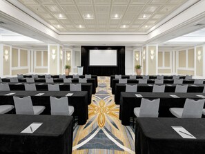 A conference room with rows of tables and chairs, a large screen at the front, and a patterned carpet.