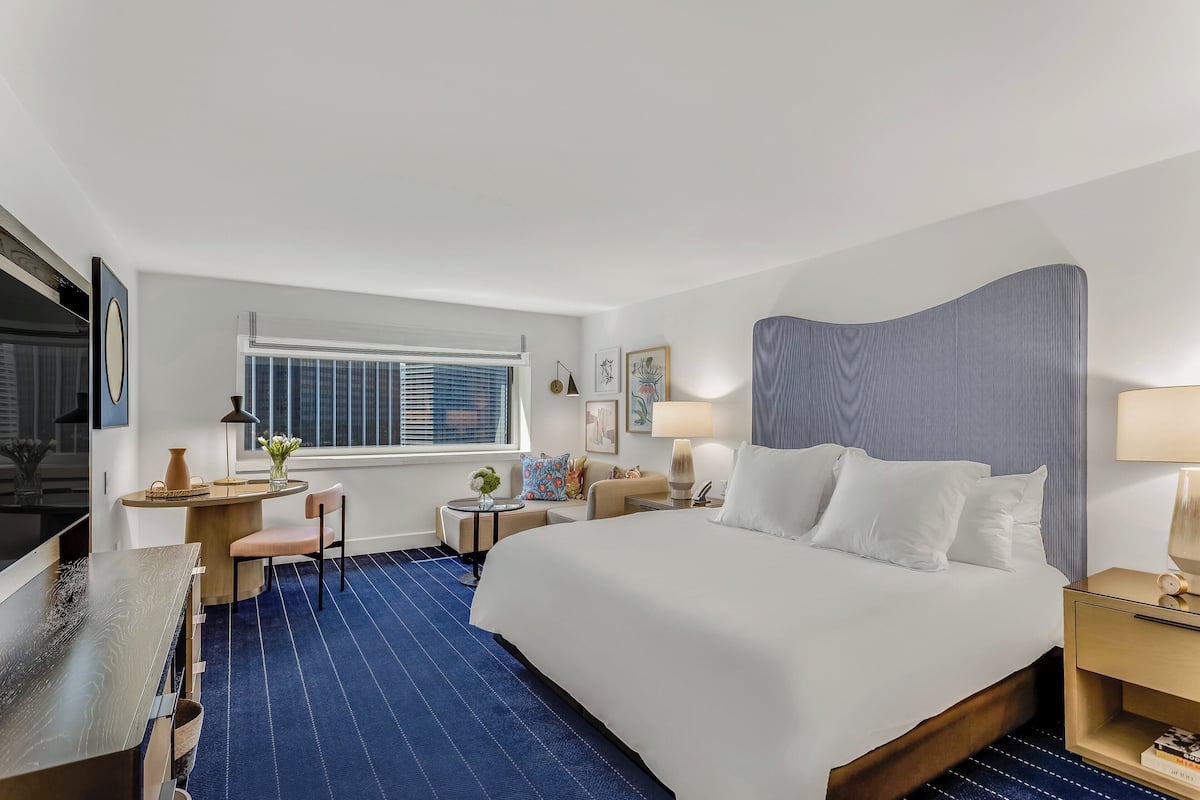 A hotel room with a large bed, a desk, a chair, a small table with a vase, and a window with blinds.