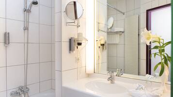 A modern bathroom with a white sink, a handheld showerhead, a mirror, and a vase with a white flower.