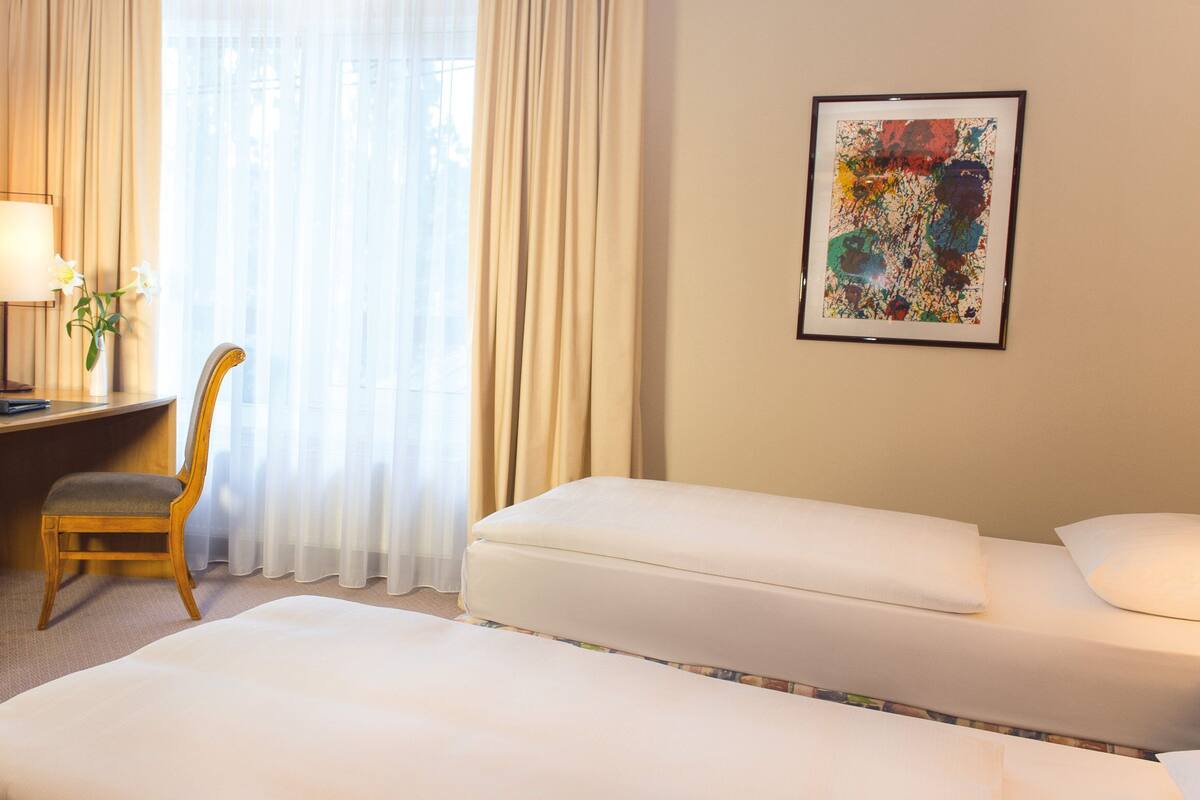 Une chambre d’h ôtel équipée d’un bureau, d’une chaise, de deux lits, d’une lampe, d’un vase avec des fleurs et d’un tableau abstrait encadré accroché au mur.