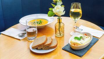 A dining table with a bowl of soup, slices of bread, a glass of water, a glass of white wine, and a vase with a white rose.