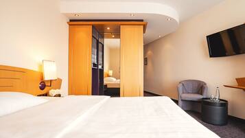 A hotel room with a large bed, a chair, a TV mounted on the wall, and a small table with a bottle and glass.