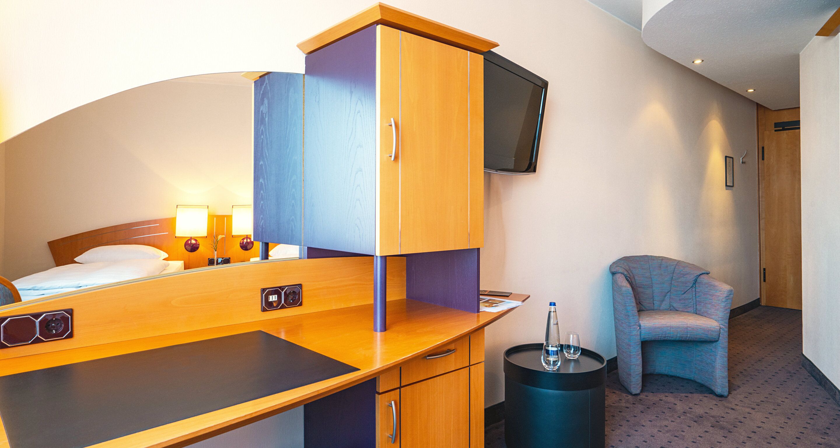 A hotel room with a bed, a desk with a computer, a chair, and a TV mounted on the wall.