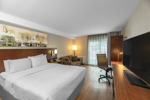 A hotel room with a large bed, a desk with a flat-screen TV, a sofa, a chair, and a painting of a cityscape on the wall. - Comfort Inn Hamilton - Stoney Creek (Hamilton)