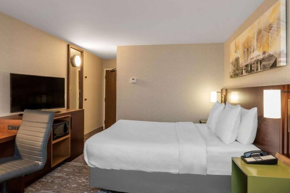 Quarto de hotel com cama, escrivaninha com cadeira, televisão, telefone e um quadro na parede.