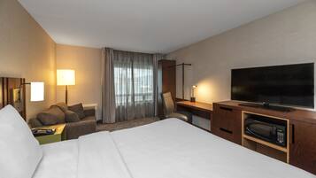 Habitación de hotel con cama, escritorio, televisor de pantalla plana, microondas, silla, mesita de noche y lámpara.