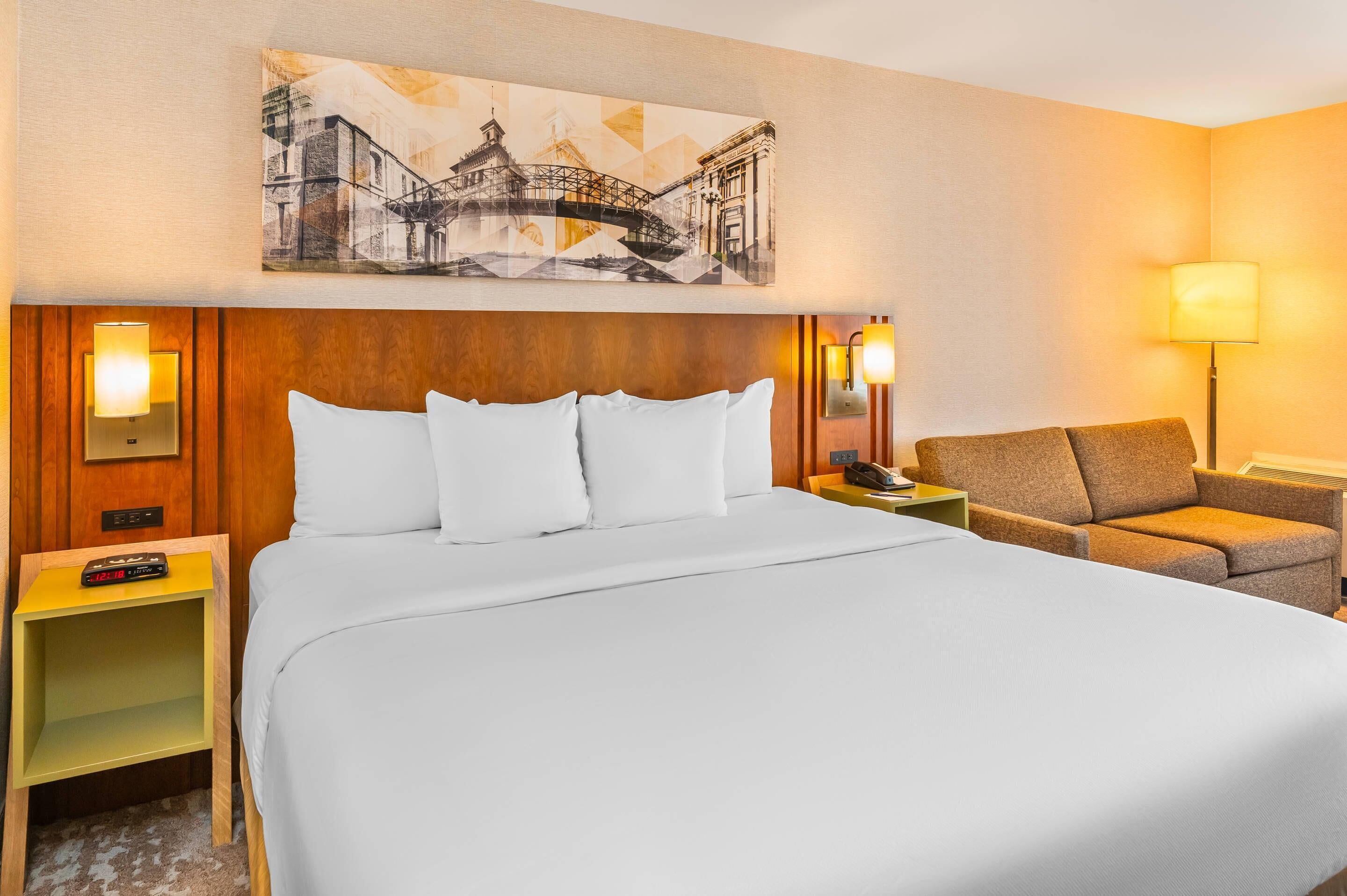 Habitación de hotel con una cama grande, una silla, una mesita de noche con lámpara y una obra de arte en la pared.