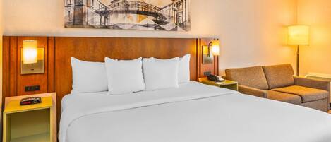 Habitación de hotel con una cama grande, una silla, una mesita de noche con lámpara y una obra de arte en la pared.