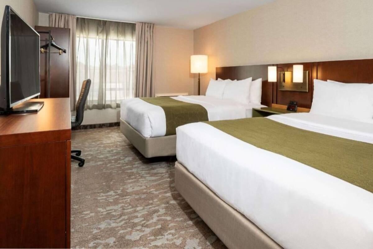 Quarto de hotel com duas camas, uma escrivaninha com televisão, uma cadeira, um abajur e uma janela com cortinas.