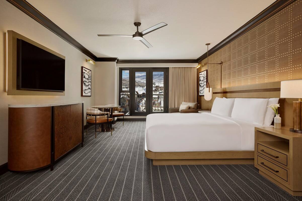 A hotel room with a bed, a desk, a chair, a TV, a ceiling fan, and a view of the outdoors.