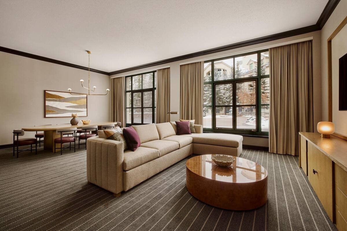 A modern living room with a sectional sofa, a dining table with chairs, a round coffee table, and large windows with curtains.