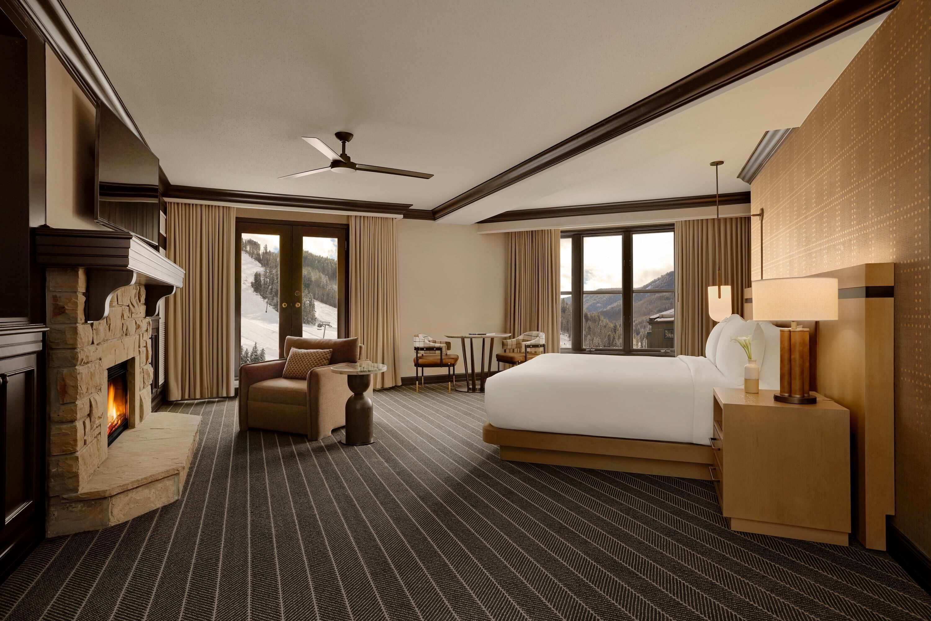 A hotel room with a fireplace, a bed, a desk, and a view of the mountains.