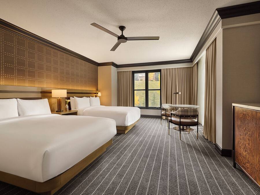 Une chambre d’hôtel avec deux lits, un ventilateur de plafond, une grande fenêtre avec des rideaux, un bureau avec une chaise et une télévision.