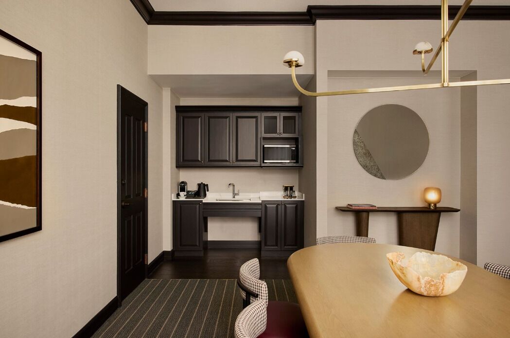 Una habitación de hotel moderna con zona de comedor, cocina con armarios oscuros, espejo redondo y una lámpara de techo original.