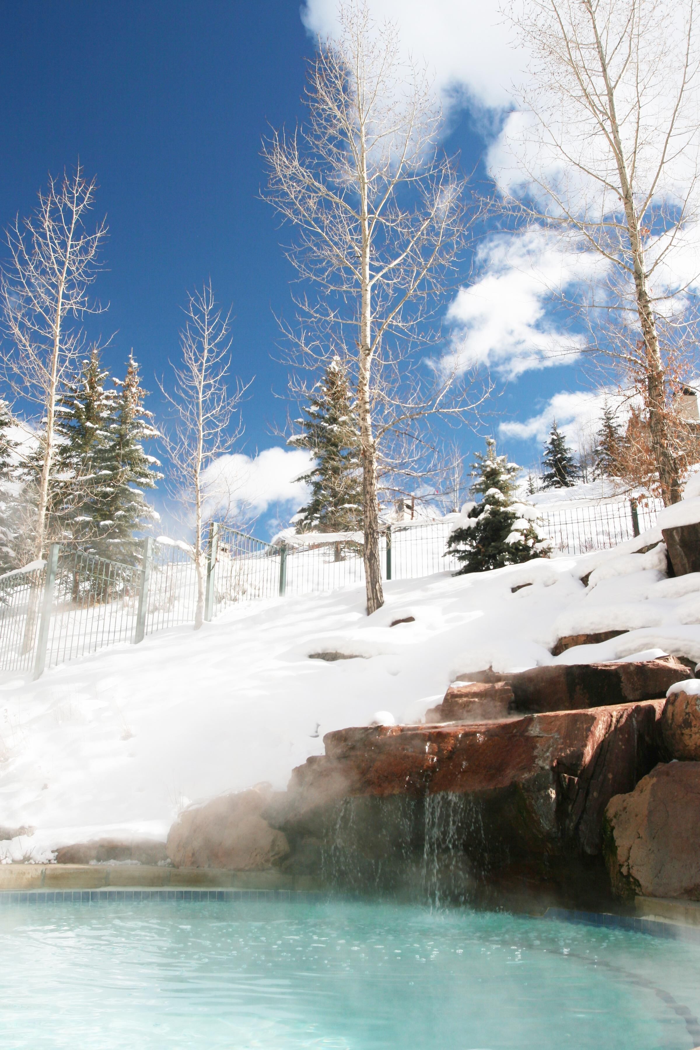 A snow-covered landscape with a clear sky, a pool with a waterfall, and snow-covered trees.