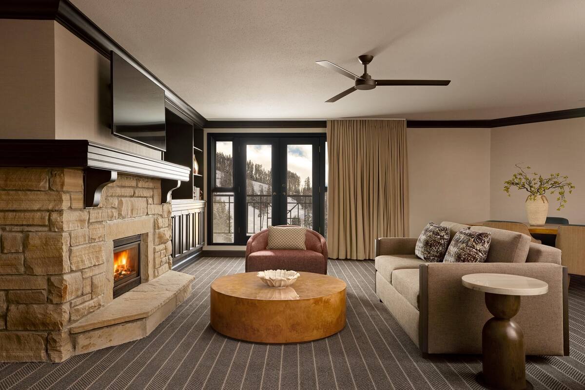 A living room with a stone fireplace, a round coffee table, a sofa, a TV mounted on the wall, and a ceiling fan.