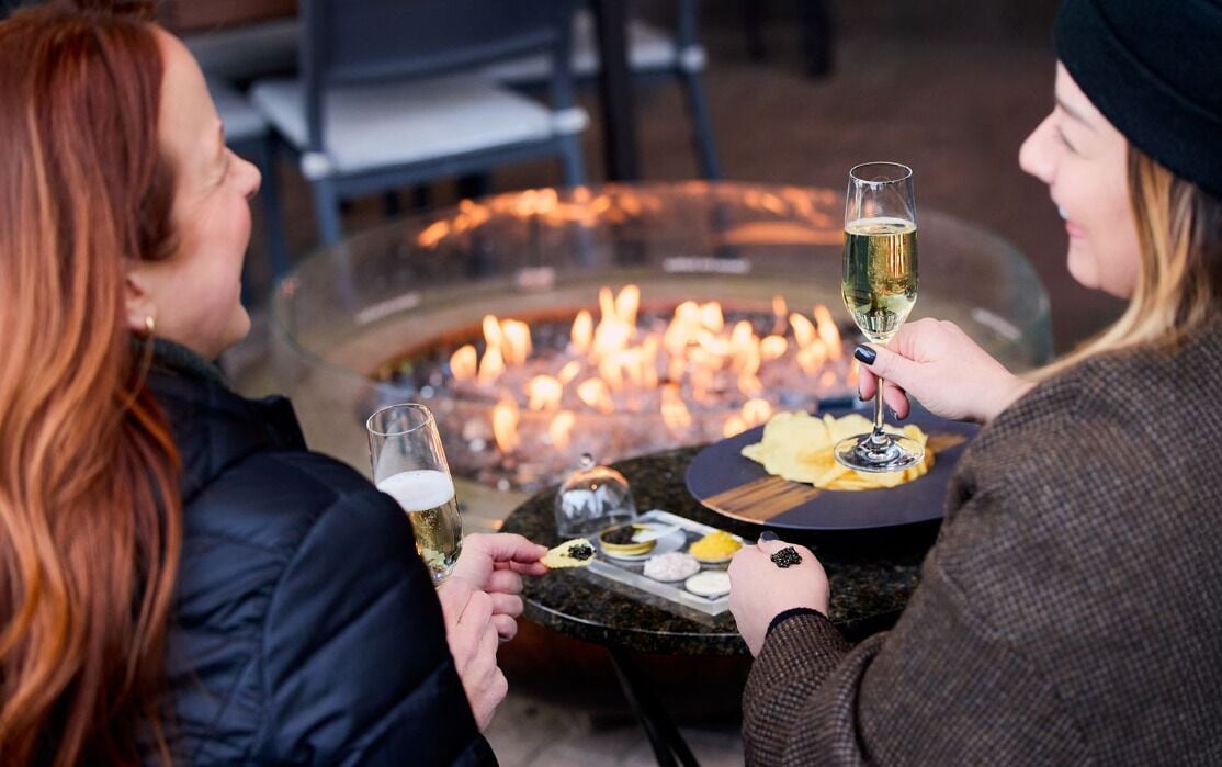 Two people sitting at a table with a glass of wine, a fire pit, and various snacks.
