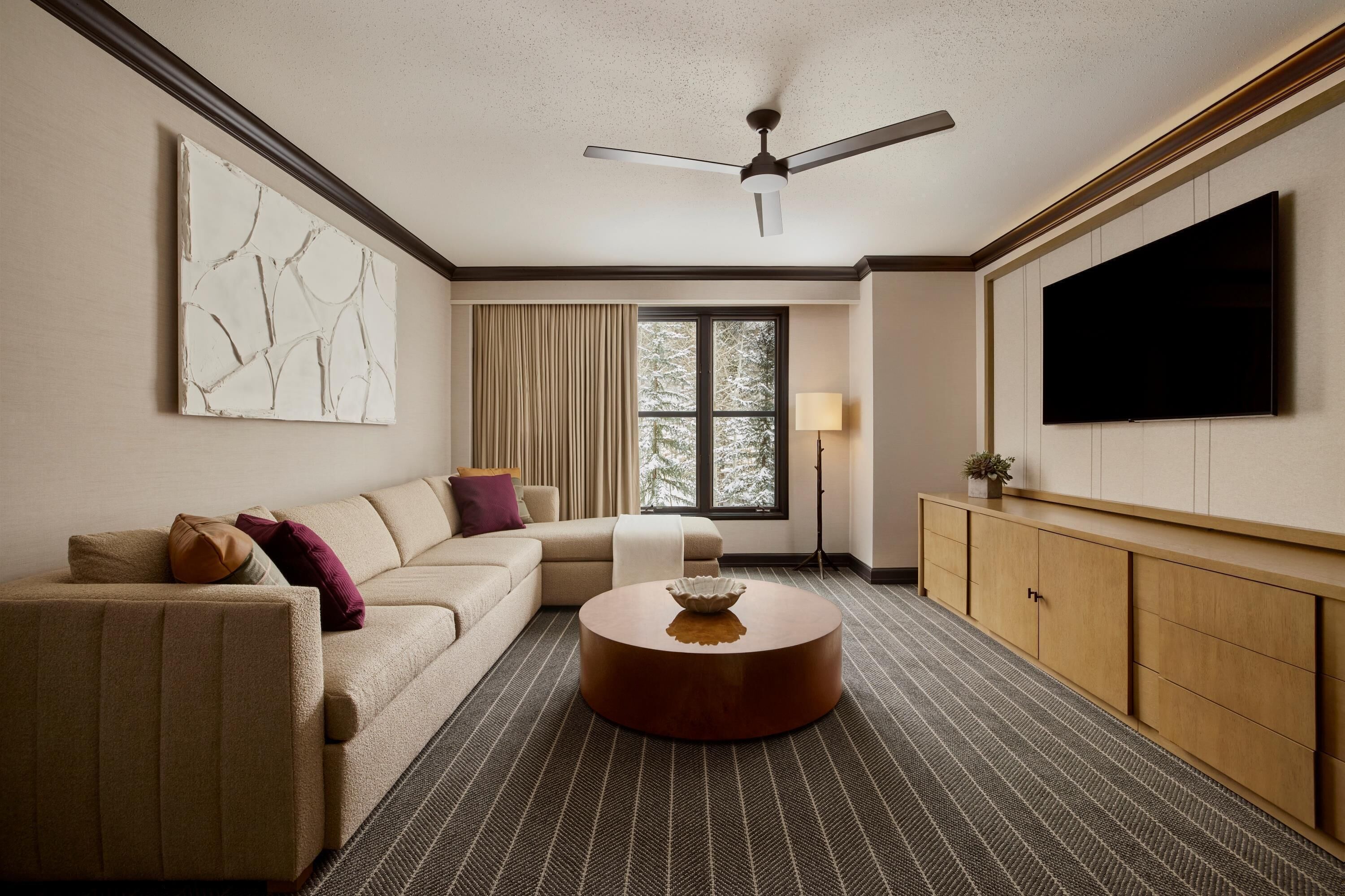 Quarto de hotel com sofá, mesa de centro redonda, televisão na parede e ventoinha no teto.