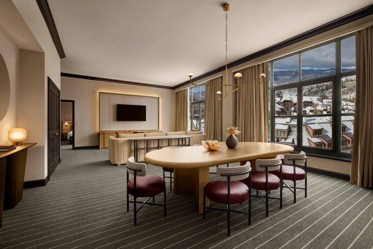 A hotel room with a large window, a dining area with a table and chairs, a sofa, and a flat-screen TV mounted on the wall.