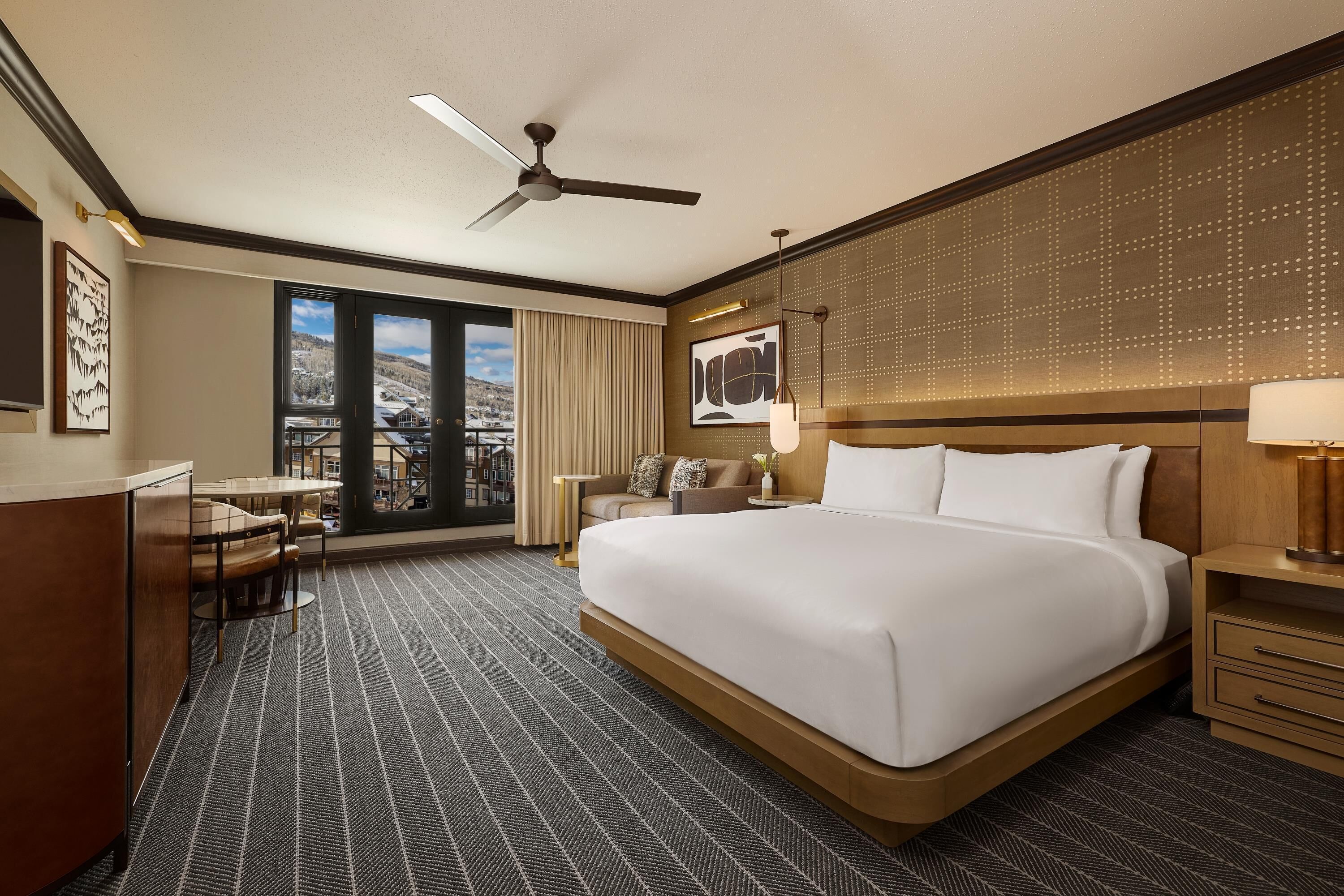 A hotel room with a large bed, a desk with chairs, a balcony with a view, and a ceiling fan.