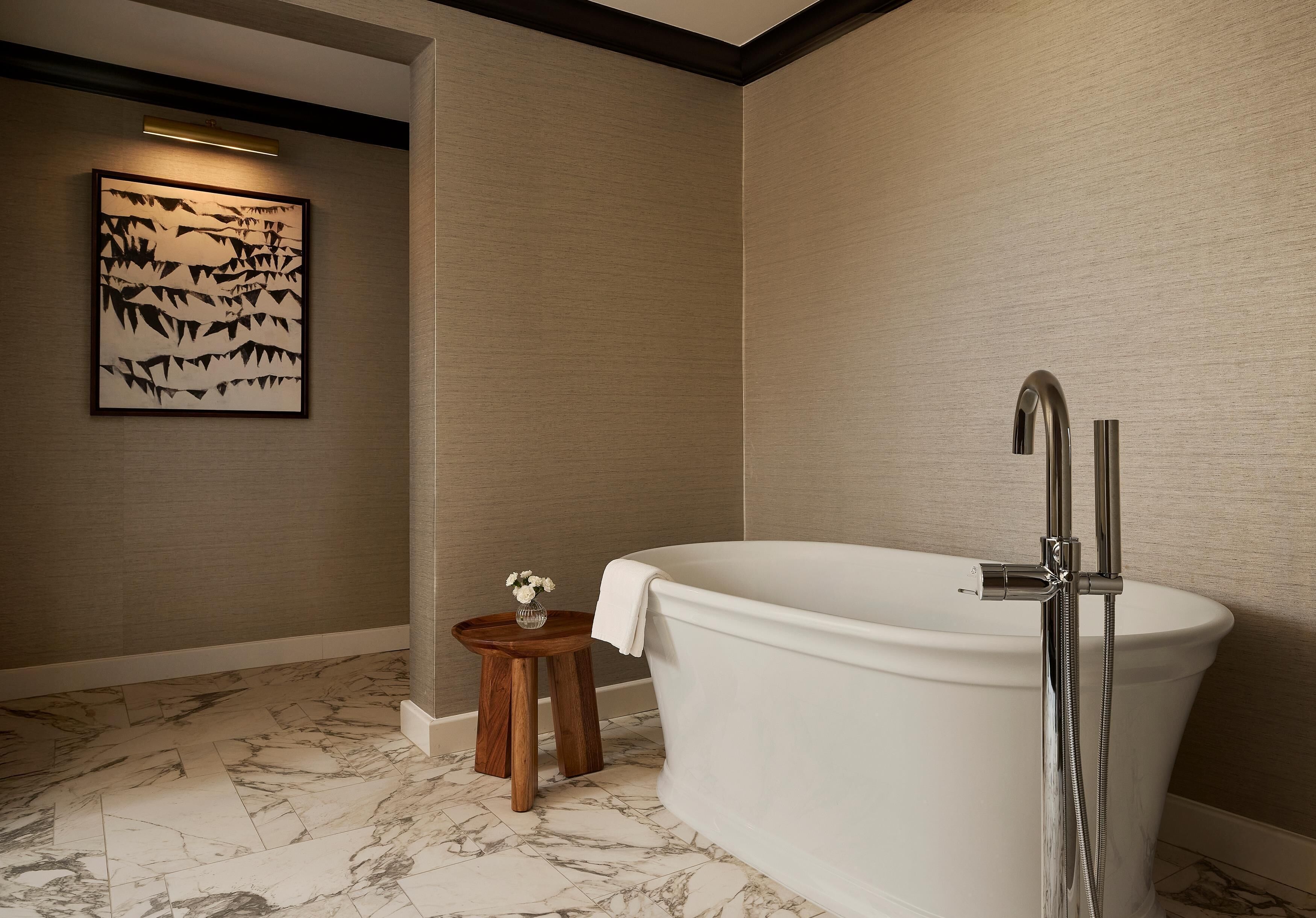 Ein Badezimmer mit freistehender Badewanne, einem kleinen hölzernen Hocker mit einer Vase und einem gerahmten Kunstwerk an der Wand.