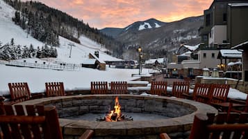 A snow-covered outdoor seating area with a stone fire pit, wooden chairs, and a view of a mountain range.