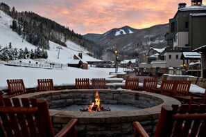 A snow-covered outdoor seating area with a stone fire pit, wooden chairs, and a view of a mountain range.