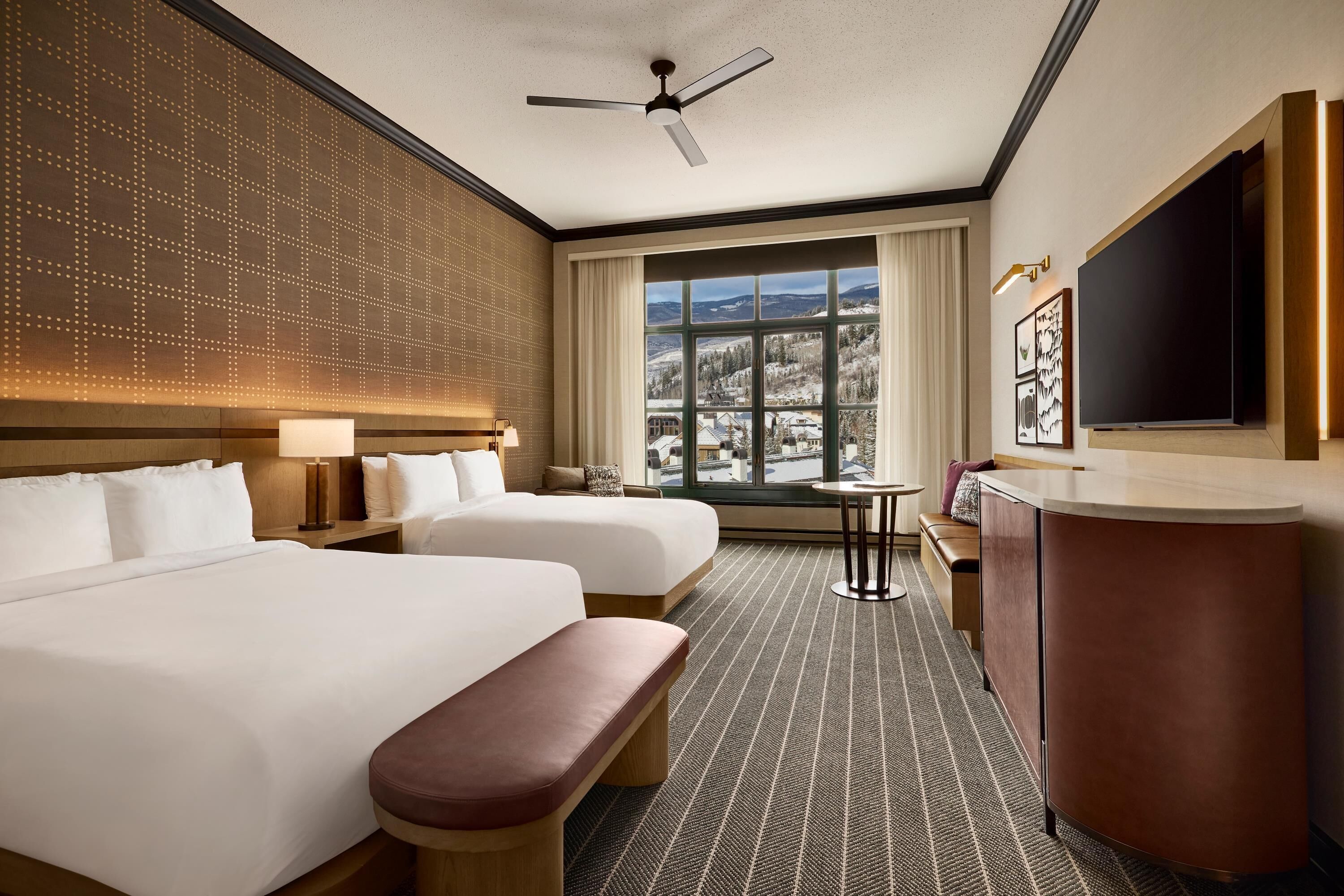 A hotel room with two beds, a desk, a TV, and a window with a view of snow-covered hills.