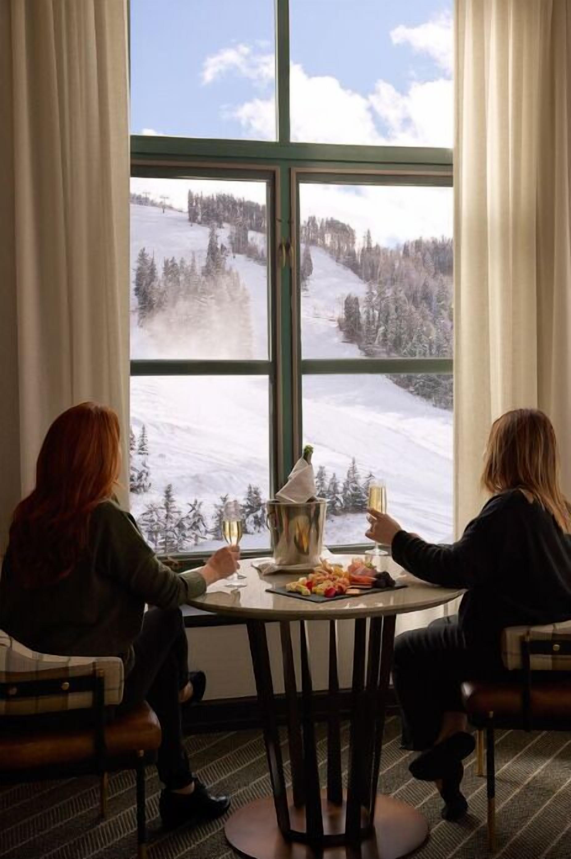Deux personnes sont assises autour d’une table ronde, devant une grande fenêtre qui donne sur un paysage enneigé.