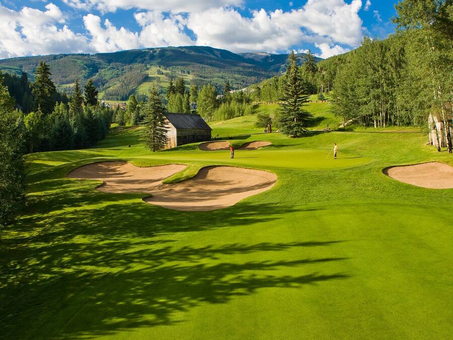 Un campo de golf con búnkeres, un club de campo y montañas al fondo.