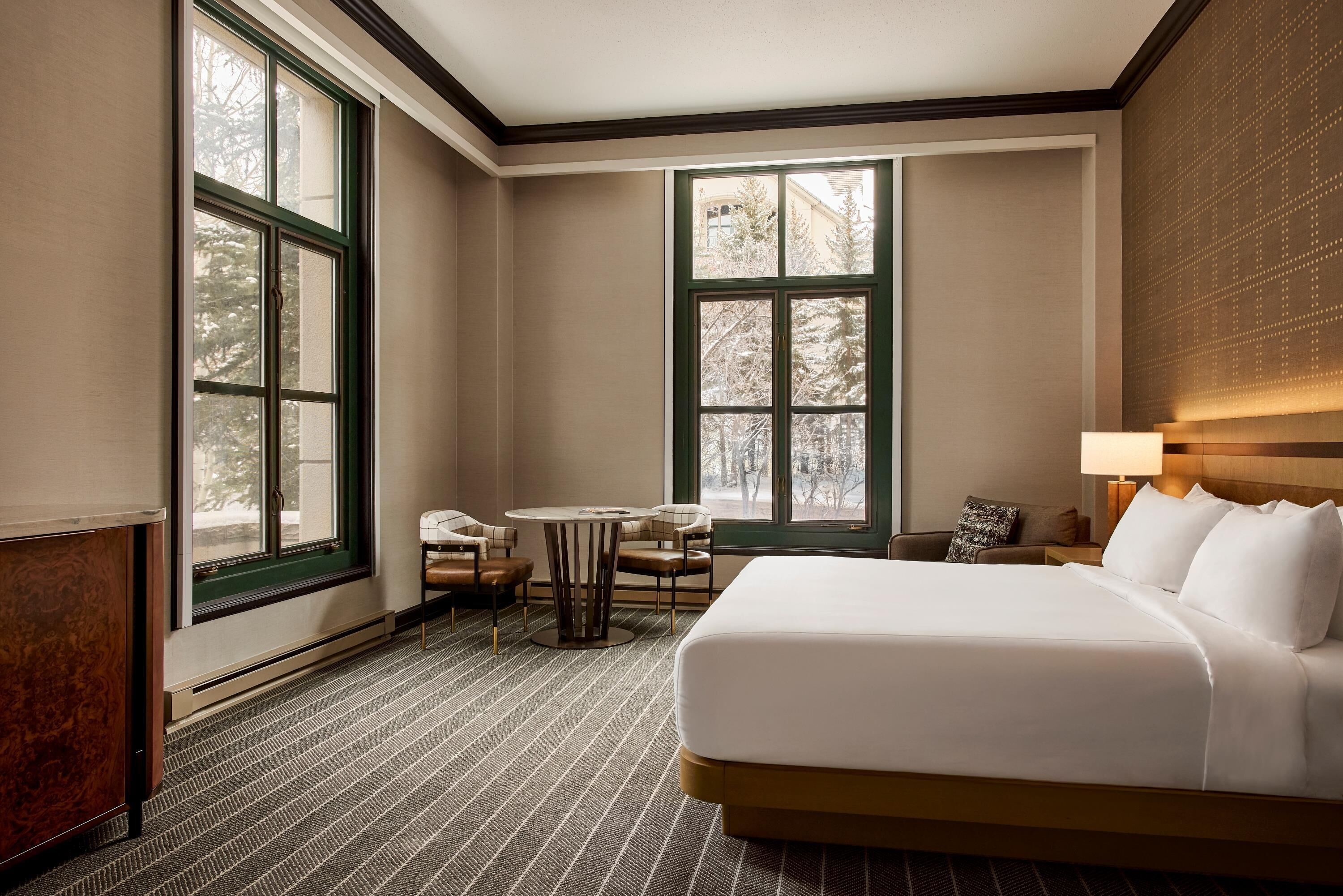 Quarto de hotel com uma cama grande, uma mesa pequena com cadeiras, um sofá e duas janelas amplas.