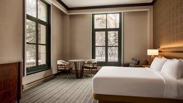 Quarto de hotel com uma cama grande, uma mesa pequena com cadeiras, um sofá e duas janelas amplas.