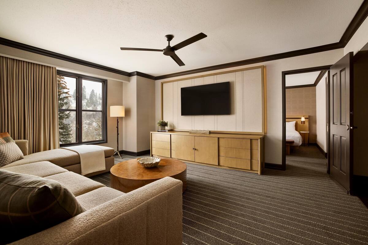 A hotel room with a sofa, a TV mounted on the wall, a ceiling fan, and a view of trees outside.